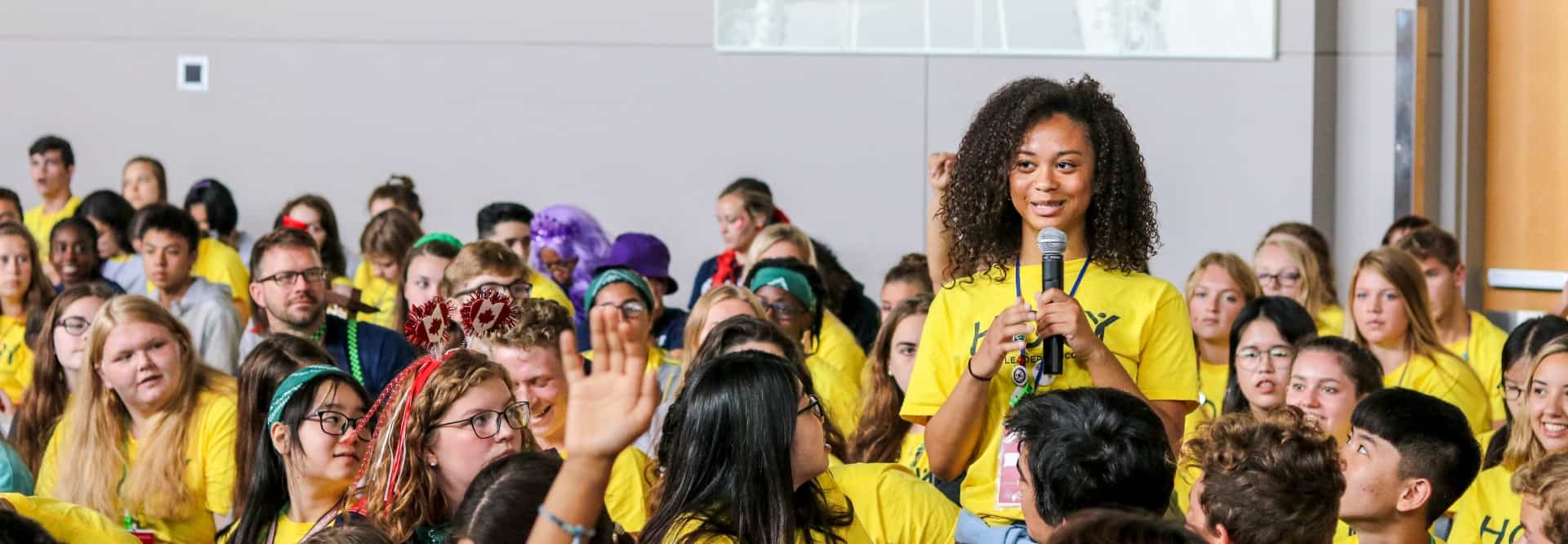 A smiling, young Black girl standing up with a microphone in a room full of HOBY students.
